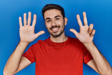 Foto de Joven hombre hispano con barba vistiendo camiseta roja sobre fondo azul mostrando y señalando con los dedos número ocho mientras sonríe confiado y feliz. - Imagen libre de derechos