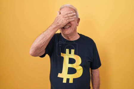 Téléchargez les photos : Homme âgé aux cheveux gris portant Bitcoin t-shirt souriant et riant avec la main sur le visage couvrant les yeux pour la surprise. concept aveugle. - en image libre de droit