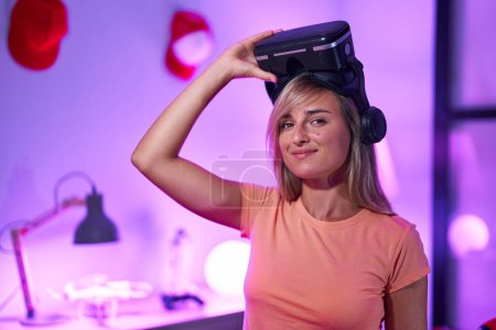 Téléchargez les photos : Jeune femme blonde streamer souriant confiant portant des lunettes de réalité virtuelle à la salle de jeux - en image libre de droit
