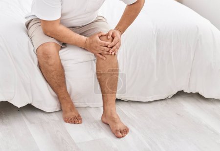 Téléchargez les photos : Jeune homme latino souffrant d'une blessure au genou assis sur le lit dans la chambre - en image libre de droit