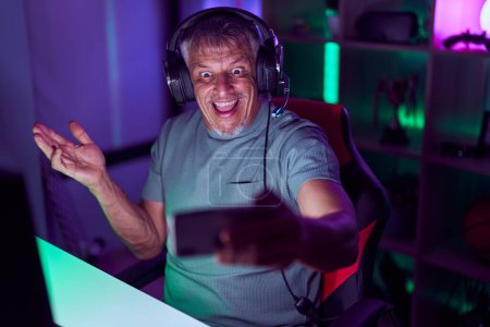 Foto de Hombre hispano con pelo gris jugando videojuegos con smartphone celebrando logros con sonrisa feliz y expresión ganadora con la mano levantada - Imagen libre de derechos