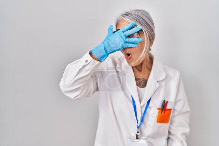 Photo for Middle age woman with grey hair wearing scientist robe peeking in shock covering face and eyes with hand, looking through fingers with embarrassed expression. - Royalty Free Image