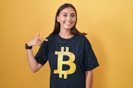 Foto de Mujer hispana joven vistiendo camiseta bitcoin mirando confiado con sonrisa en la cara, señalándose con los dedos orgullosos y felices. - Imagen libre de derechos