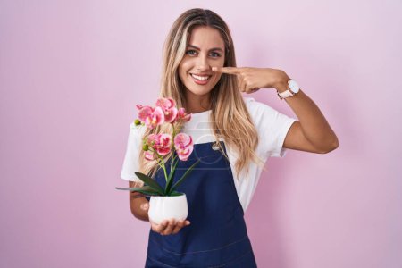 Foto de Mujer rubia joven con delantal de jardinero sosteniendo la planta señalando con el dedo a la mano y la nariz, sonriendo alegre. concepto de belleza - Imagen libre de derechos