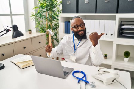 Téléchargez les photos : Afro-Américain travaillant à la clinique médicale tenant marteau pointant le pouce vers le côté souriant heureux avec la bouche ouverte - en image libre de droit