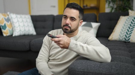 Photo for Young hispanic man watching tv sitting on floor with boring expression at home - Royalty Free Image