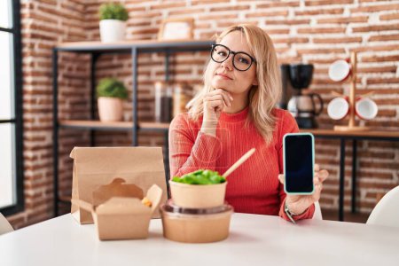 Foto de Mujer rubia comiendo comida para llevar mostrando la pantalla del teléfono inteligente cara seria pensando en la pregunta con la mano en la barbilla, reflexivo acerca de la idea confusa - Imagen libre de derechos