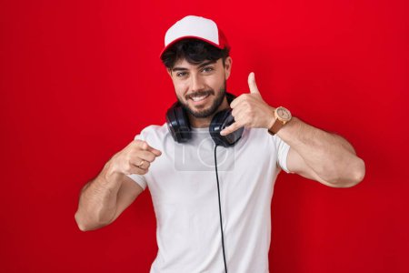 Foto de Hombre hispano con barba con sombrero de gamer y auriculares sonriendo hablando por teléfono y señalándote. Llámame.. - Imagen libre de derechos