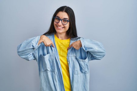 Foto de Mujer hispana joven de pie sobre fondo azul mirando confiado con sonrisa en la cara, señalándose con los dedos orgullosos y felices. - Imagen libre de derechos