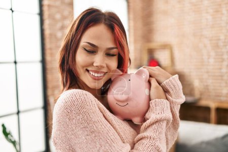 Foto de Young caucasian woman smiling confident hugging piggy bank at new home - Imagen libre de derechos