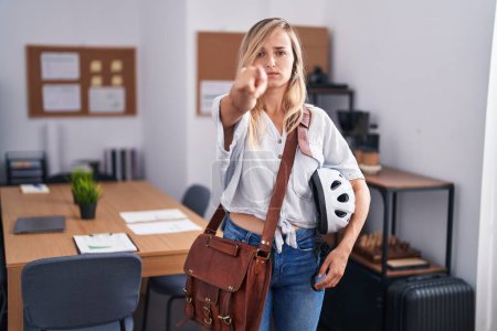 Téléchargez les photos : Jeune femme blonde travaillant au bureau tenant un casque de vélo pointant du doigt la caméra et vers vous, geste confiant et sérieux - en image libre de droit
