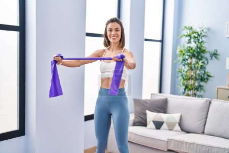 Photo for Young beautiful hispanic woman smiling confident using elastic band training at home - Royalty Free Image