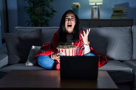 Photo for Hispanic woman eating popcorn watching a movie on the sofa crazy and mad shouting and yelling with aggressive expression and arms raised. frustration concept. - Royalty Free Image