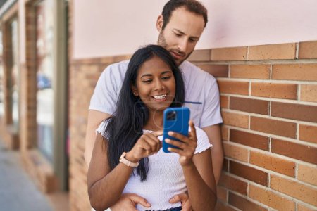 Téléchargez les photos : Man and woman interracial couple hugging each other using smartphone at street - en image libre de droit