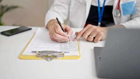 Téléchargez les photos : Homme d'âge moyen avec médecin des cheveux gris en utilisant l'écriture d'ordinateur portable sur le presse-papiers à la clinique - en image libre de droit