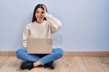 Foto de Mujer joven usando el ordenador portátil sentado en el suelo en casa haciendo buen gesto con la mano sonriendo, ojos mirando a través de los dedos con la cara feliz. - Imagen libre de derechos