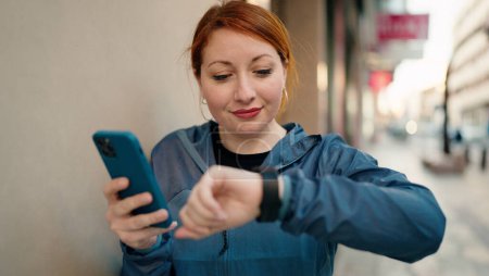 Photo for Young redhead woman wearing sportswear using smartphone looking stopwatch at street - Royalty Free Image