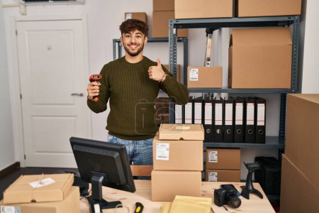 Téléchargez les photos : Homme arabe avec barbe travaillant dans le commerce des petites entreprises tenant scanner de code à barres souriant heureux et positif, pouce levé faire excellent et signe d'approbation - en image libre de droit