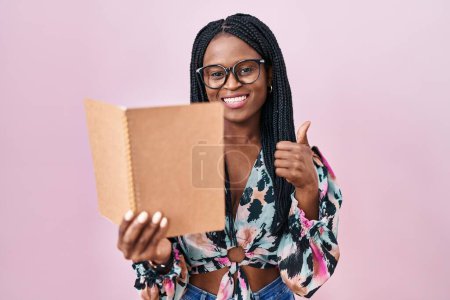 Photo for African woman with braids reading a book smiling happy and positive, thumb up doing excellent and approval sign - Royalty Free Image