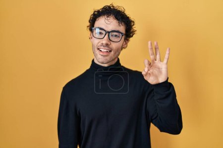 Foto de Hombre hispano de pie sobre fondo amarillo mostrando y señalando hacia arriba con los dedos número tres mientras sonríe confiado y feliz. - Imagen libre de derechos