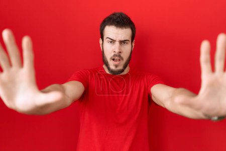 Téléchargez les photos : Jeune homme hispanique portant un t-shirt rouge décontracté faisant un geste d'arrêt avec les paumes des mains, expression de colère et de frustration - en image libre de droit