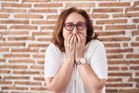 Photo for Senior woman with glasses standing over bricks wall laughing and embarrassed giggle covering mouth with hands, gossip and scandal concept - Royalty Free Image