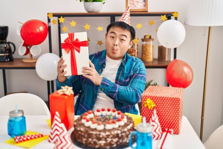 Téléchargez les photos : Jeune homme chinois célébrant l'anniversaire avec gâteau et cadeau faisant visage de poisson avec la bouche et les yeux plissés, fou et comique. - en image libre de droit