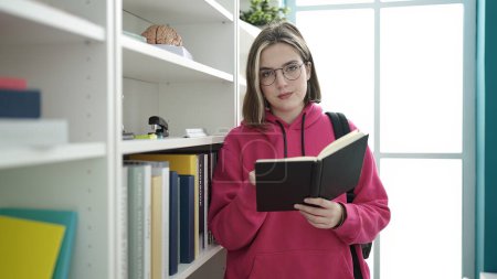 Téléchargez les photos : Young blonde woman student reading book standing at library university - en image libre de droit