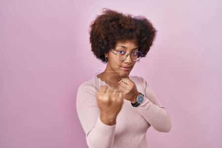 Téléchargez les photos : Jeune femme afro-américaine debout sur fond rose prêt à se battre avec geste de défense poing, visage en colère et contrarié, peur du problème - en image libre de droit