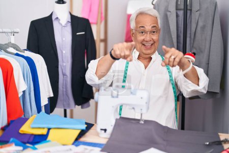 Foto de Hombre de mediana edad con modista de pelo gris usando máquina de coser que apunta a usted y la cámara con los dedos, sonriendo positivo y alegre - Imagen libre de derechos