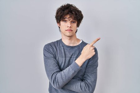 Téléchargez les photos : Jeune homme debout sur fond isolé pointant avec le doigt de la main sur le côté montrant la publicité, visage sérieux et calme - en image libre de droit