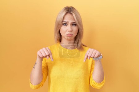 Foto de Mujer caucásica joven con suéter amarillo apuntando hacia abajo mirando triste y molesto, indicando la dirección con los dedos, infeliz y deprimido. - Imagen libre de derechos