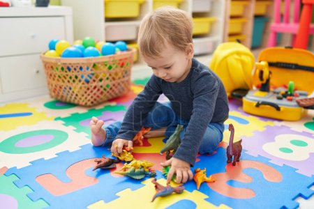 Téléchargez les photos : Adorable bambin blond jouant avec des jouets dino assis sur le sol à la maternelle - en image libre de droit