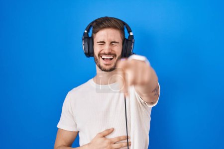 Foto de Hombre hispano con barba escuchando música usando auriculares riéndose de ti, señalando con el dedo a la cámara con la mano sobre el cuerpo, expresión de vergüenza - Imagen libre de derechos