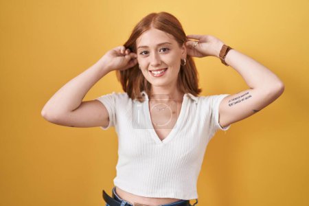 Photo for Young redhead woman standing over yellow background smiling pulling ears with fingers, funny gesture. audition problem - Royalty Free Image