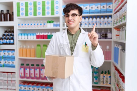 Téléchargez les photos : Jeune homme non binaire travaillant à la pharmacie drugstore tenant boîte sourire heureux pointant avec la main et le doigt vers le côté - en image libre de droit