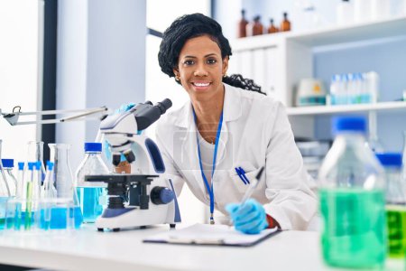 Foto de Mujer afroamericana vistiendo uniforme científico usando microscopio escribir en documento en laboratorio - Imagen libre de derechos