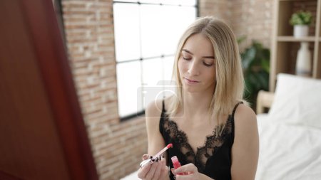 Téléchargez les photos : Jeune femme blonde mettre rouge à lèvres assis sur le lit regardant sexy au miroir dans la chambre - en image libre de droit