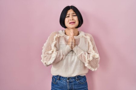 Téléchargez les photos : Jeune femme asiatique debout sur fond rose suppliant et priant avec les mains ainsi que l'expression d'espoir sur le visage très émotionnel et inquiet. mendier. - en image libre de droit