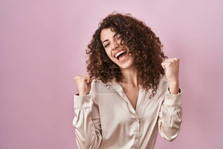 Téléchargez les photos : Femme hispanique avec les cheveux bouclés debout sur fond rose célébrant surpris et étonnés pour le succès avec les bras levés et les yeux fermés. concept gagnant. - en image libre de droit