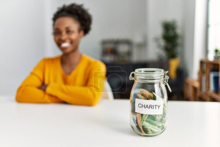 Foto de Mujer afroamericana sentada en mesa sosteniendo frasco de caridad con billetes de rand en casa - Imagen libre de derechos