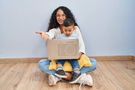 Téléchargez les photos : Jeune hispanique mère et enfant en utilisant ordinateur portable assis sur le sol souriant joyeuse offrant main de paume donnant assistance et acceptation. - en image libre de droit