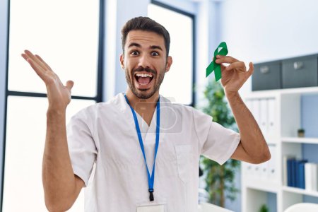 Photo for Young hispanic doctor man holding support green ribbon at the clinic celebrating victory with happy smile and winner expression with raised hands - Royalty Free Image