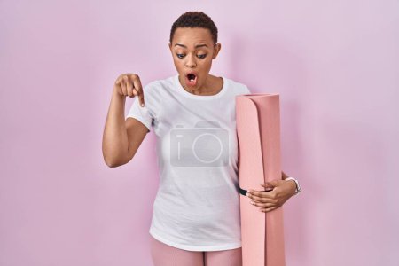 Téléchargez les photos : Belle femme afro-américaine tenant tapis de yoga pointant vers le bas avec les doigts montrant la publicité, visage surpris et bouche ouverte - en image libre de droit
