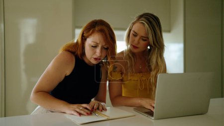 Foto de Two women using laptop writing on notebook sitting on table at kitchen - Imagen libre de derechos