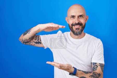 Foto de Hombre hispano con tatuajes de pie sobre fondo azul haciendo gestos con las manos mostrando un signo de tamaño grande y grande, símbolo de medida. sonriendo mirando a la cámara. concepto de medición. - Imagen libre de derechos