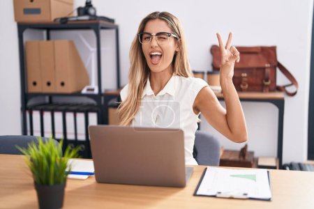 Young blonde woman working at the office wearing glasses smiling with happy face winking at the camera doing victory sign. number two.  mug #648245508