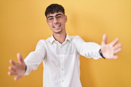 Photo for Young hispanic man standing over yellow background looking at the camera smiling with open arms for hug. cheerful expression embracing happiness. - Royalty Free Image