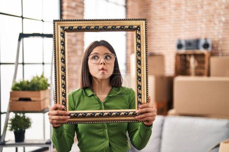 Photo for Young hispanic woman at new home holding empty frame making fish face with mouth and squinting eyes, crazy and comical. - Royalty Free Image