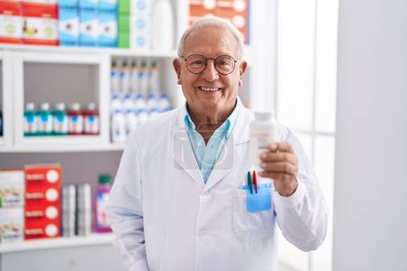 Foto de Hombre mayor de pelo gris farmacéutico sonriendo confiado sosteniendo pastillas botella en la farmacia - Imagen libre de derechos